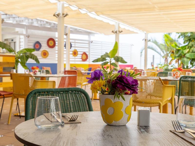 Colorful terrace with table set and flowers in yellow vase under sunshade.