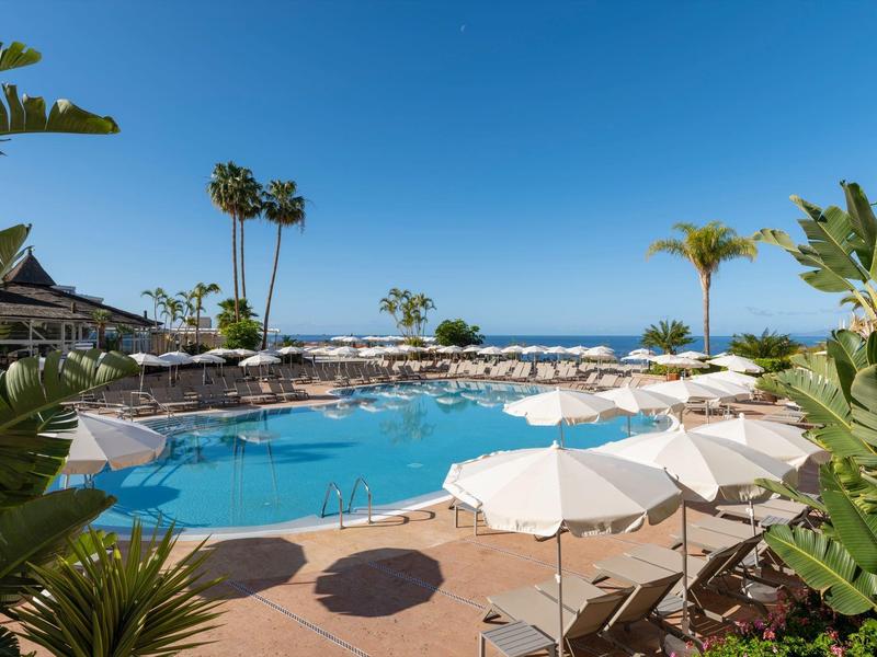 View of a pool with umbrellas and palm trees against a blue sky and sea view.