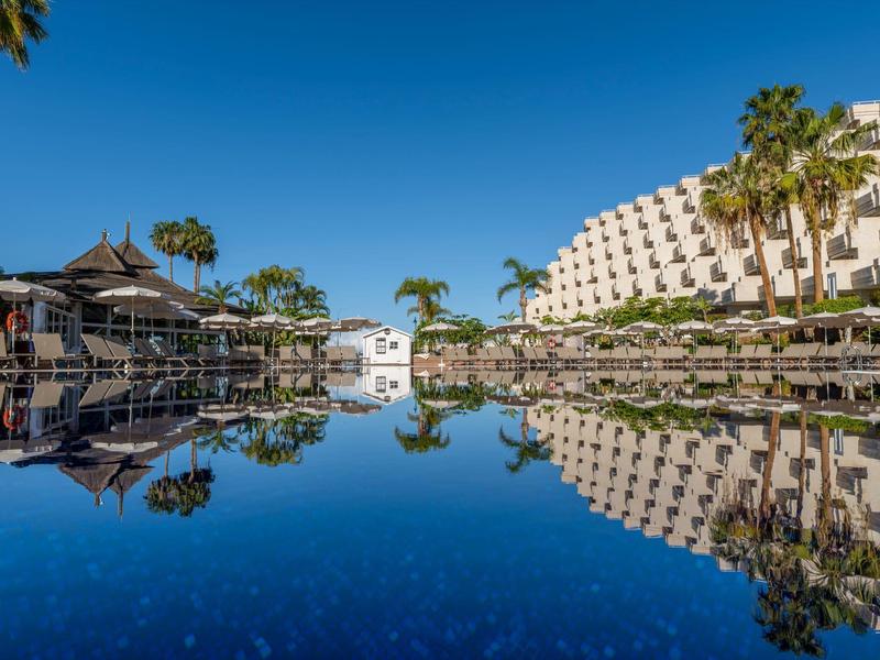 Calm pool reflecting palm trees and hotels under clear daytime sky.