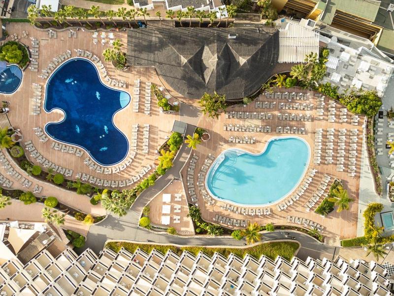 Aerial view of two swimming pools with sun loungers surrounded by palm trees at a hotel resort.