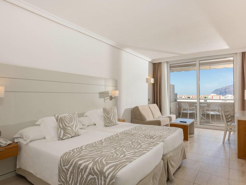 Bright hotel room with two beds, seating area, and balcony overlooking the sea.