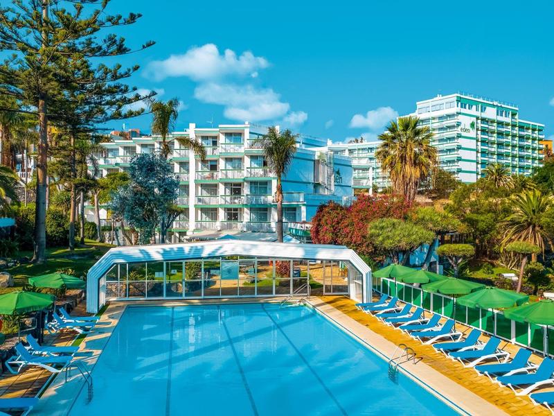 Leisure pool with sun loungers surrounded by palm trees and white hotel building under blue sky