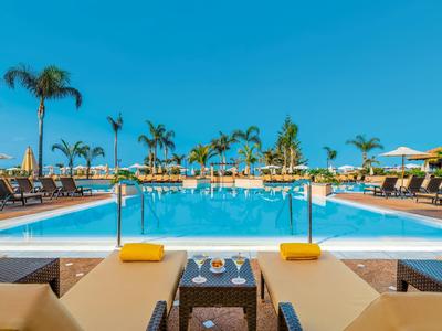 Grand piscine extérieure avec transats, palmiers et ciel bleu clair dans un complexe hôtelier.