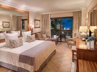 Chambre d'hôtel confortable avec grand lit, fauteuils et balcon donnant sur des palmiers la nuit.