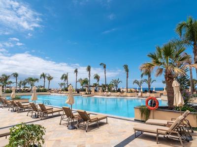 Grande piscine d'hôtel avec palmiers et chaises longues sous un ciel bleu clair.