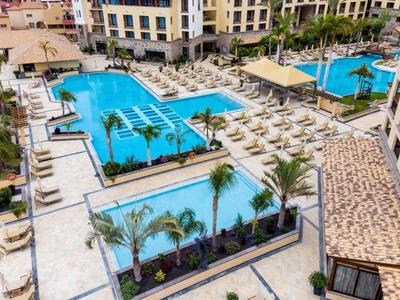 Grande piscine d'hôtel avec chaises longues et palmiers, entourée de bâtiments à plusieurs étages.