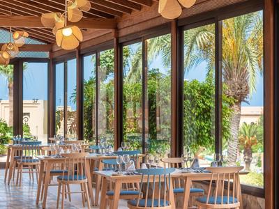 Restaurant lumineux avec mobilier en bois et grandes fenêtres donnant sur un jardin tropical.