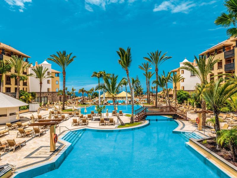 Piscine d'hôtel avec eau bleue, chaises longues et palmiers sous un ciel clair.