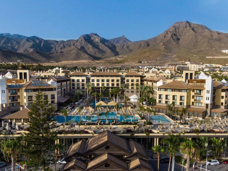 Hôtel de luxe avec grande piscine, entouré de montagnes et de palmiers par temps ensoleillé.