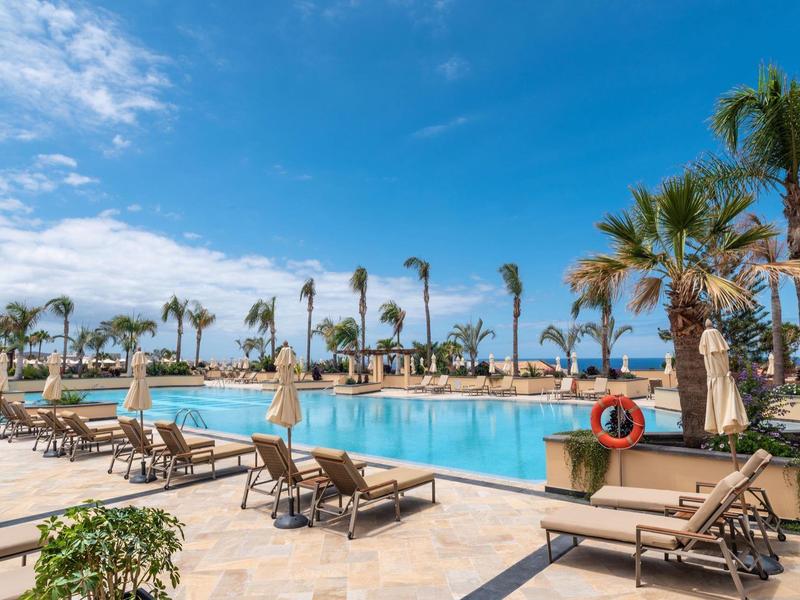 Grande piscine d'hôtel avec palmiers et chaises longues sous un ciel bleu clair.