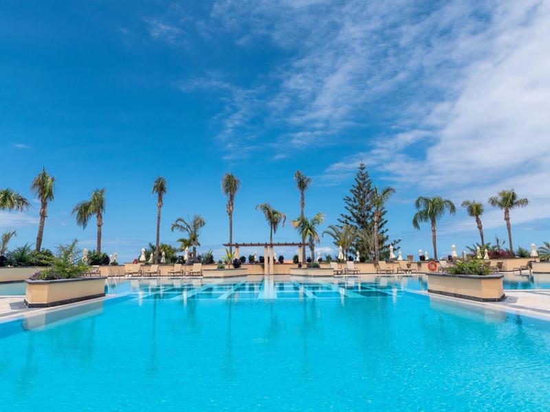 Grande piscine extérieure avec parasols et palmiers sous un ciel bleu dans une station balnéaire.