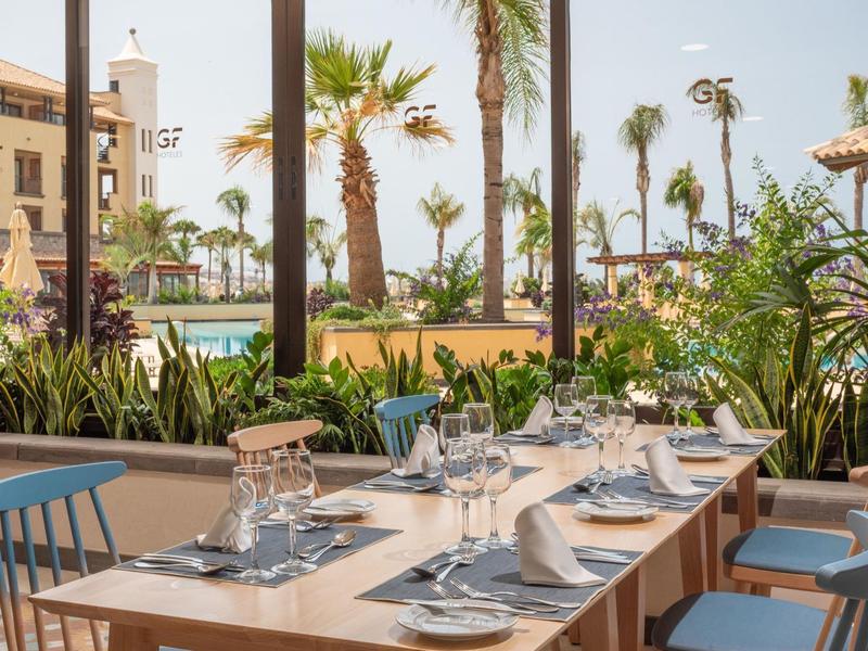 Table dressée avec verres et assiettes à côté de grandes fenêtres donnant sur des palmiers et une terrasse extérieure.