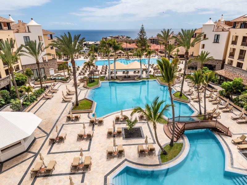 Grand piscine d'hôtel entourée de transats, palmiers et bâtiments avec vue sur la mer.