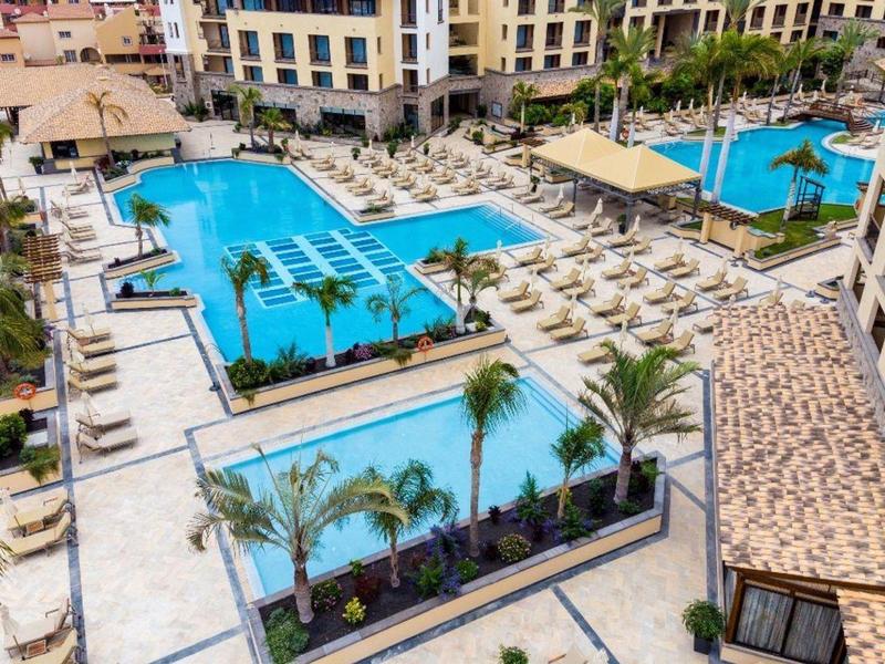 Grande piscine d'hôtel avec chaises longues et palmiers, entourée de bâtiments à plusieurs étages.
