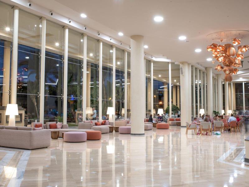 Lobby d'hôtel lumineuse avec grand lustre, sièges confortables et fenêtres du sol au plafond.