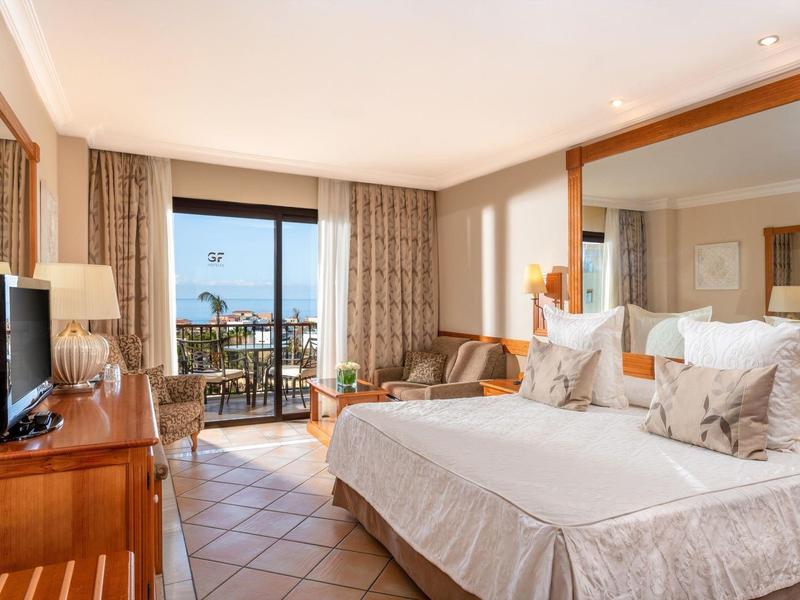 Chambre d'hôtel lumineuse avec lit double, TV, canapé et balcon avec vue sur la mer.