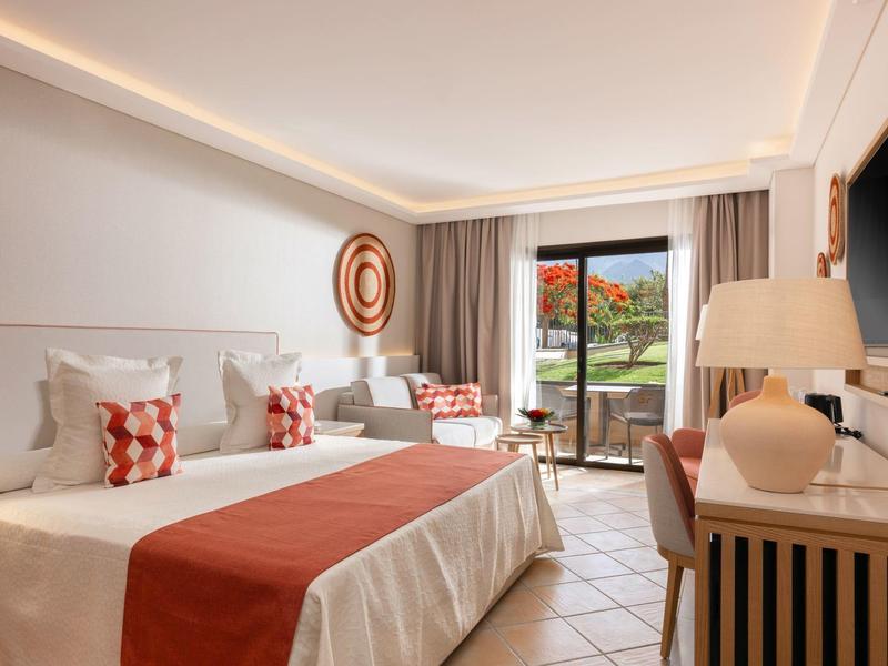 Chambre d'hôtel lumineuse avec deux lits, accents rouges et vue sur terrasse fleurie.