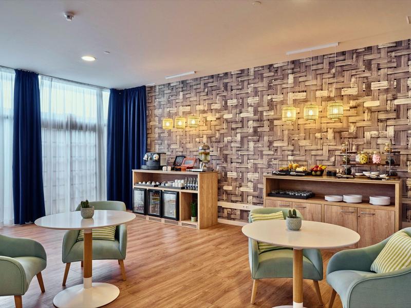 Moderner Frühstücksraum mit Holzboden, gemusteter Wand, runden Tischen und grünen Sesseln.