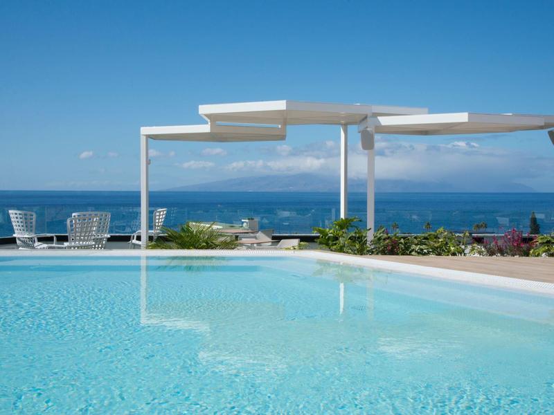 Clear pool beside white pergolas overlooking blue sea and sky at a sunny resort.