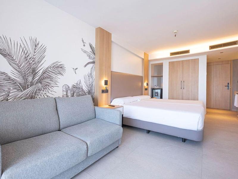 Modern hotel room with bed, grey sofa, wooden accents, and tropical wall art.