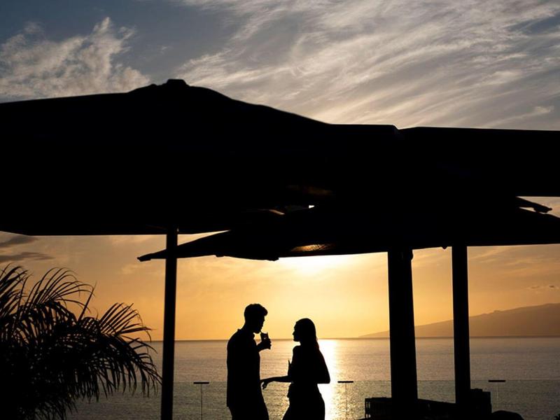 Silhouettes of two people standing under a large umbrella by the sea at sunset.