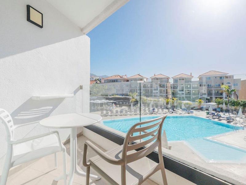Balcony with table and chairs overlooking a sunlit pool and hotel buildings under blue sky