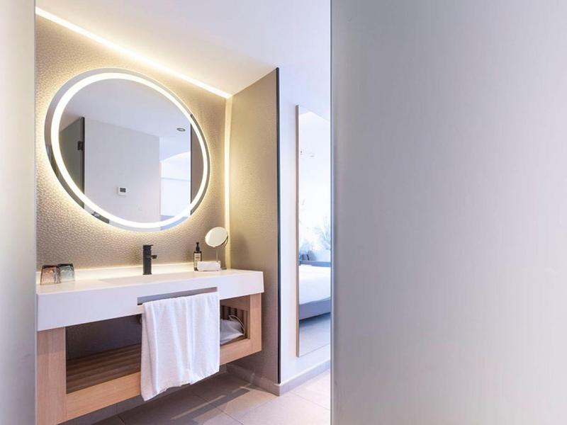 Modern hotel bathroom with a circular backlit mirror, white countertop, and wooden cabinet.