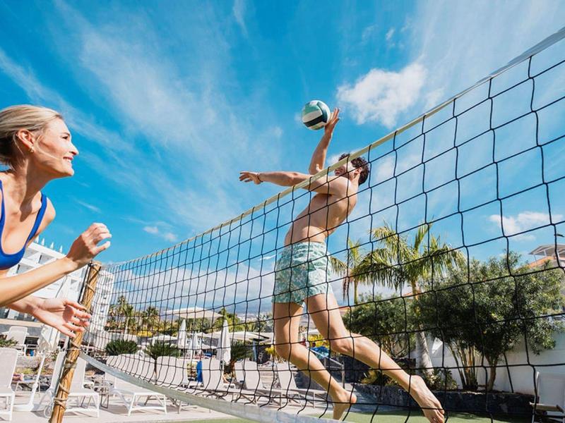 People playing volleyball outdoors on a sunny day near palm trees and buildings.