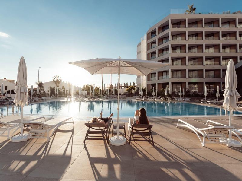 Hotel pool area with sun loungers, umbrellas, and a multi-story building in the background at sunset.