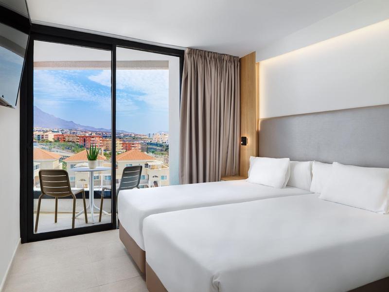 Modern hotel room with twin beds and balcony overlooking a sunny cityscape.
