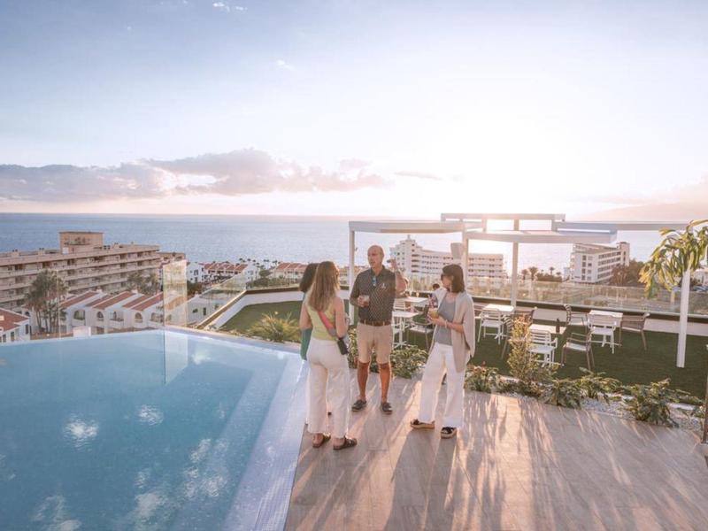 Three people enjoy a rooftop pool with city and sea views during sunset.