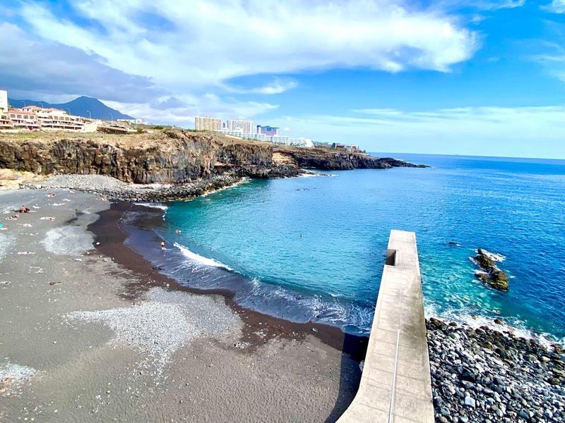 Coastal view with rocky shore, sandy beach, and a wooden pier leading into clear blue sea under a partly cloudy sky.