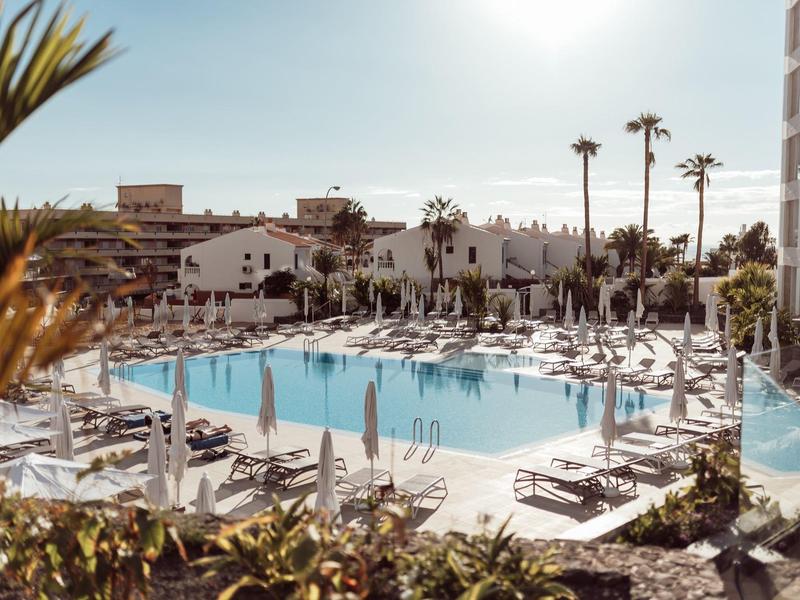 Sunny resort with a large pool surrounded by sun loungers and palm trees.