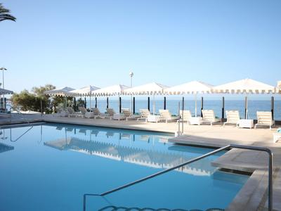 Piscina exterior moderna con tumbonas y vistas al mar bajo un cielo azul despejado.