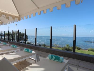 Terrasse mit Liegestühlen und Glasgeländer, Blick auf grünen Garten und blauen Ozean bei klarem Himmel.