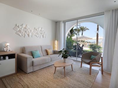 Heller Wohnraum mit grauer Couch, Holzstuhl, Pflanzen und großem Fenster mit Blick ins Freie.