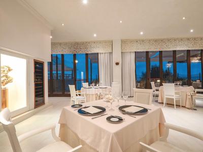 Elegantes, helles Restaurant mit weißen Möbeln, gedeckten Tischen und großen Fenstern mit Meerblick bei Nacht.