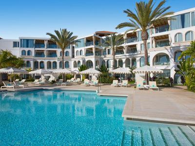 Complejo hotelero moderno con gran piscina, palmeras y tumbonas bajo un cielo azul.