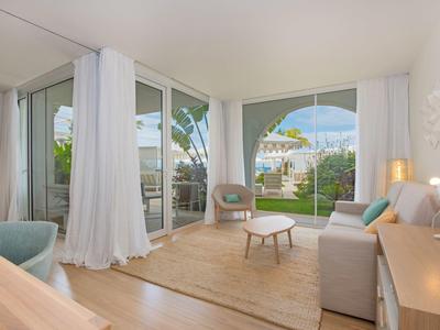 Helles Wohnzimmer mit großen Fenstern, weißen Vorhängen, Holzmöbeln und Blick auf Garten.