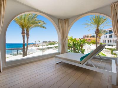 Terrasse mit Liegestuhl, Palmen und Meerblick durch zwei große runde Fensterbögen.