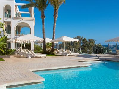 Moderner Pool mit türkisem Wasser, weißen Liegestühlen, Sonnenschirmen und Palmen unter blauem Himmel.