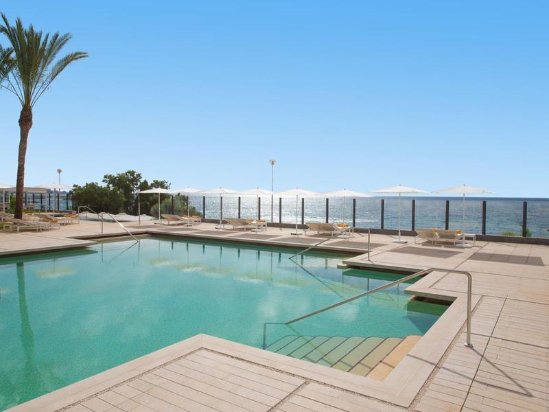 Ein klarer Pool mit Treppen, umgeben von einer Holzterrasse und Palmen am sonnigen Himmel.