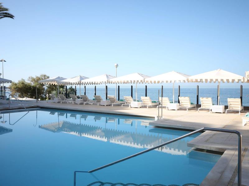 Schwimmbad mit blauer Wasserfläche, Sonnenliegen und Palmen, dahinter Meer und klarer Himmel.