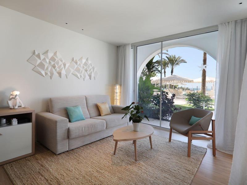 Heller Wohnraum mit grauer Couch, Holzstuhl, Pflanzen und großem Fenster mit Blick ins Freie.