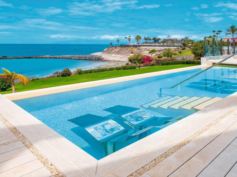 Moderner Pool mit Liegestühlen im Wasser, blauer Himmel und Meer im Hintergrund.