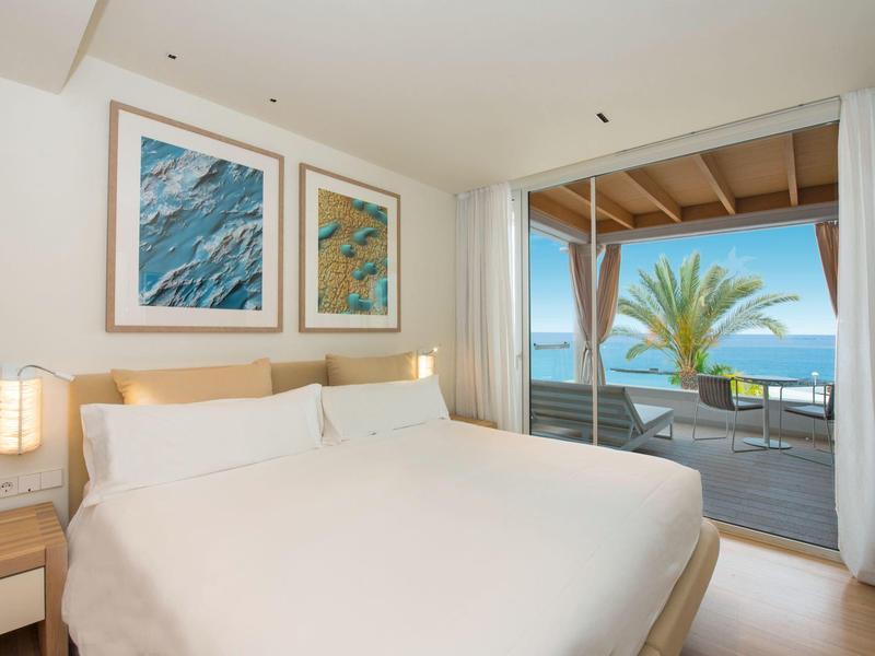 Modernes Schlafzimmer mit Doppelbett, Kunstwerken an der Wand und Balkon mit Meerblick und Palme.