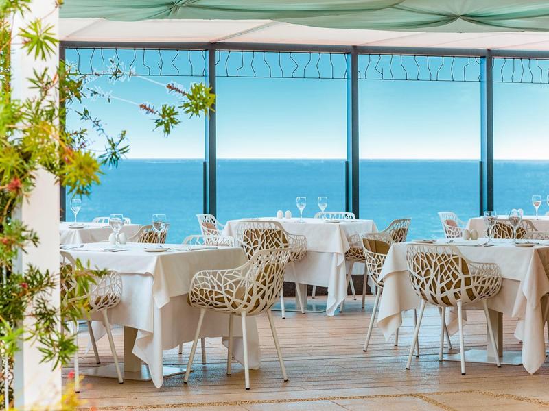 Offenes Restaurant mit weißen Tischen und dekorativen Stühlen, Blick auf blauen Ozean und Himmel.
