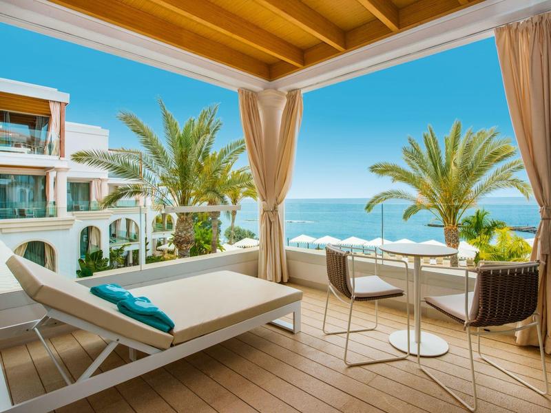 Balkon mit Holzboden, Liege, zwei Stühlen, Tisch, Palmen und Meerblick unter blauem Himmel.