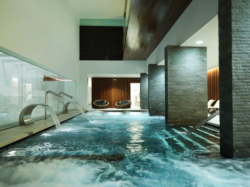 Modernes Hallenbad mit klarem Wasser, grauen Steinwänden und indirekter Beleuchtung.