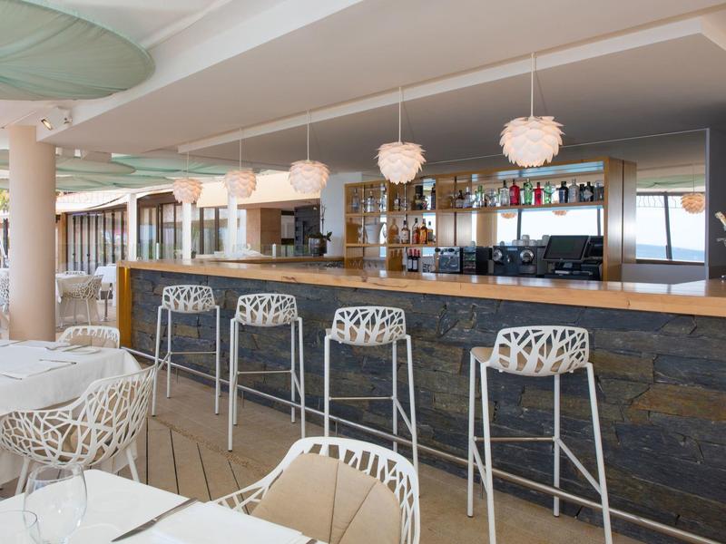 Moderner Barbereich mit fünf weißen Hockern, hängenden Lampen und offenem Regal mit Flaschen.